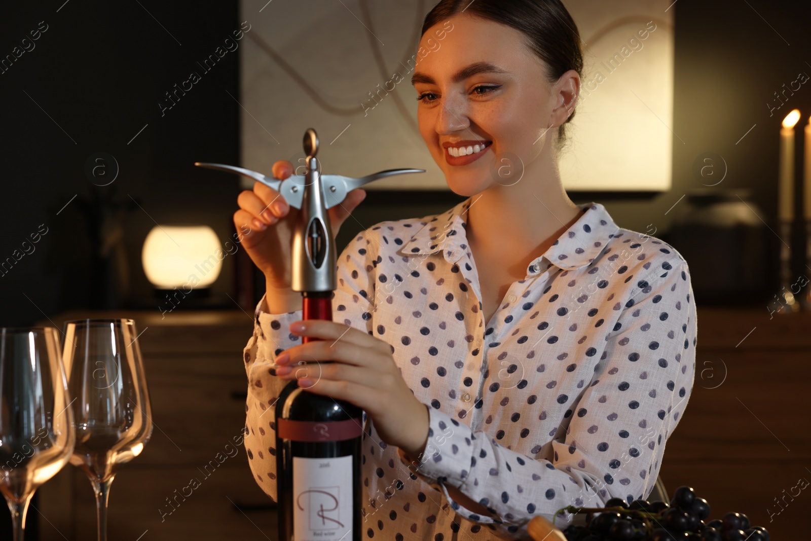 Photo of Romantic dinner. Happy woman opening wine bottle with corkscrew at home