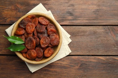 Photo of Bowl of tasty apricots and green leaves on wooden table, top view with space for text. Dried fruits