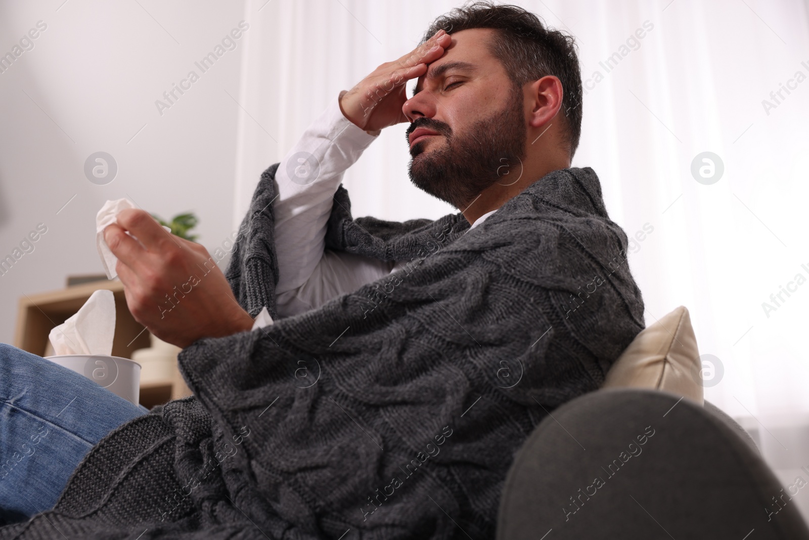 Photo of Sick man wrapped in blanket with tissue on sofa at home. Cold symptoms