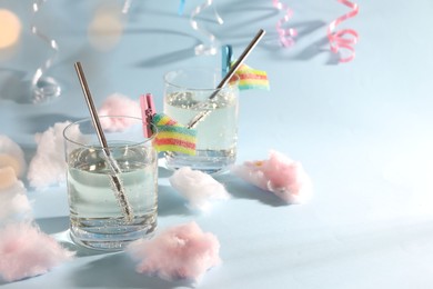 Photo of Tasty cocktails in glasses decorated with gummy candies and cotton candy on light blue background, space for text