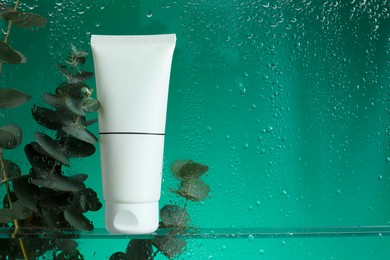 Photo of Tube with moisturizing cream and eucalyptus branches on green background, view through wet glass. Space for text