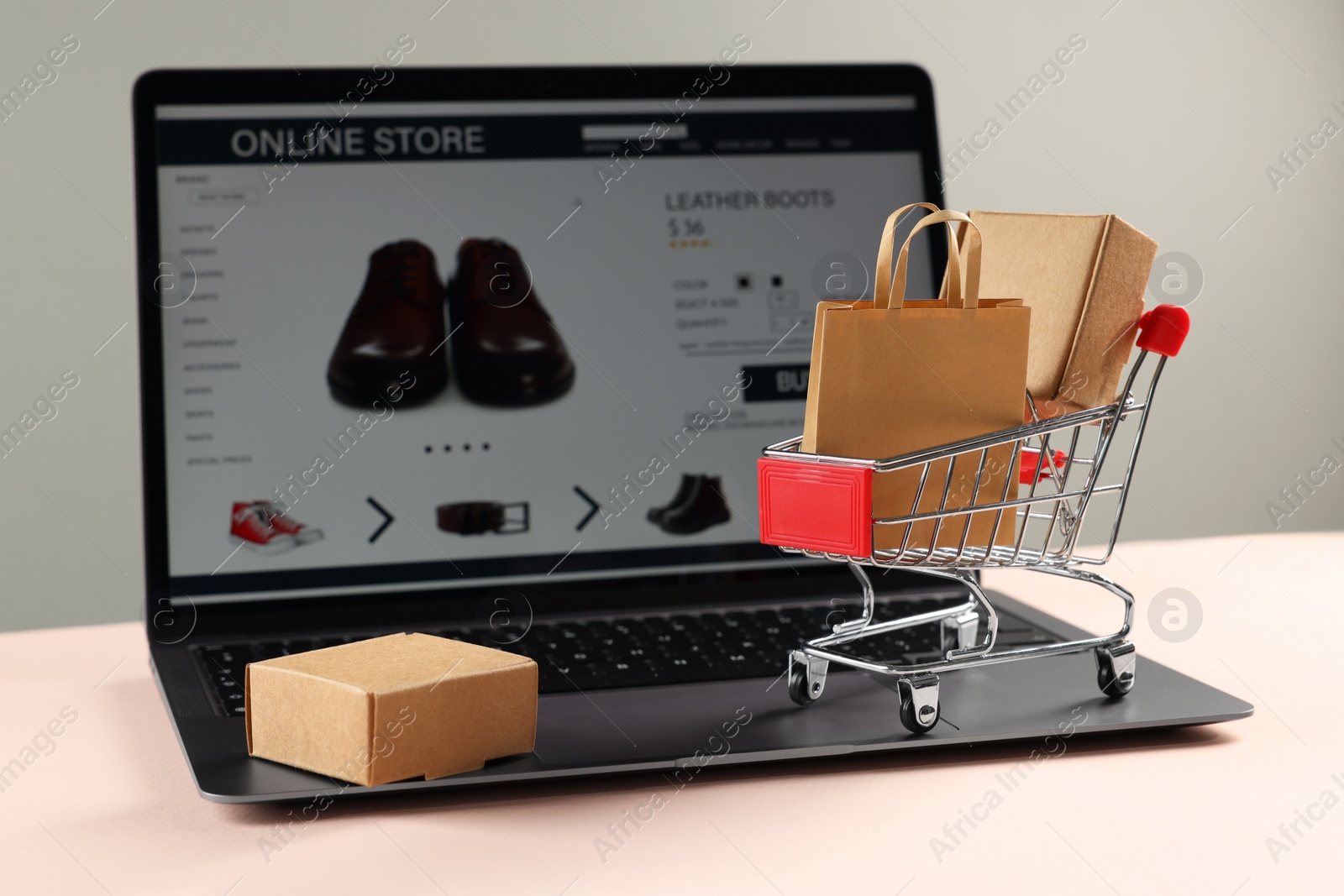 Photo of Online store. Laptop, mini shopping cart and purchases on beige table