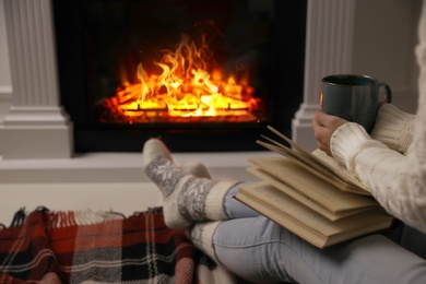 Woman with cup of hot drink and book resting near fireplace at home, closeup