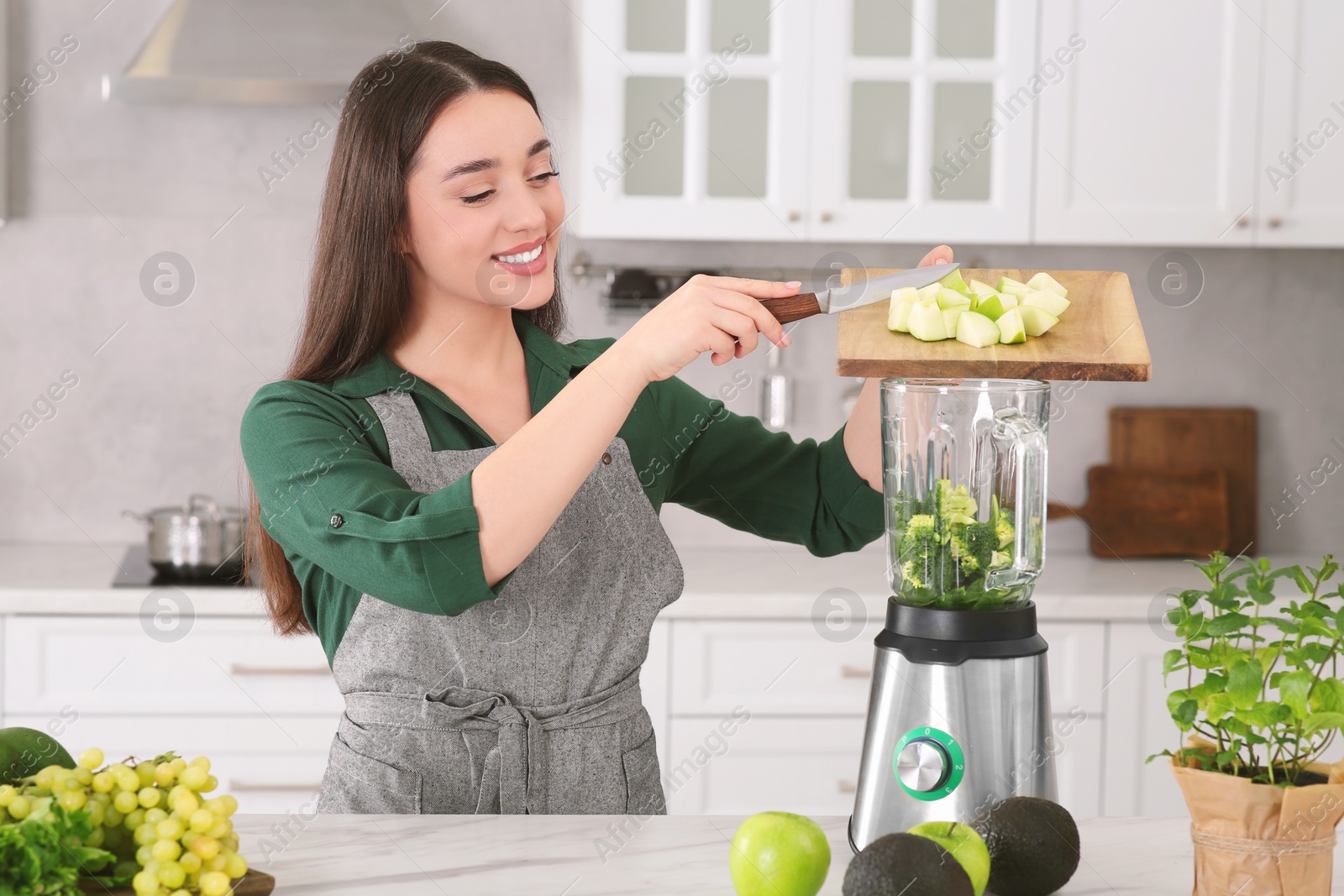 Photo of Woman adding apples into blender with ingredients for smoothie at white table in kitchen