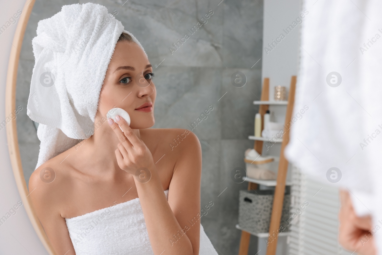 Photo of Young woman cleaning her face with cotton pad near mirror in bathroom