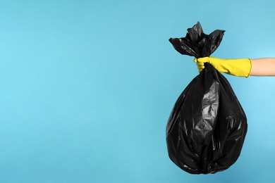 Photo of Janitor in rubber glove holding trash bag full of garbage on light blue background, closeup. Space for text