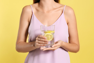Photo of Young woman with glass of lemon water on yellow background, closeup