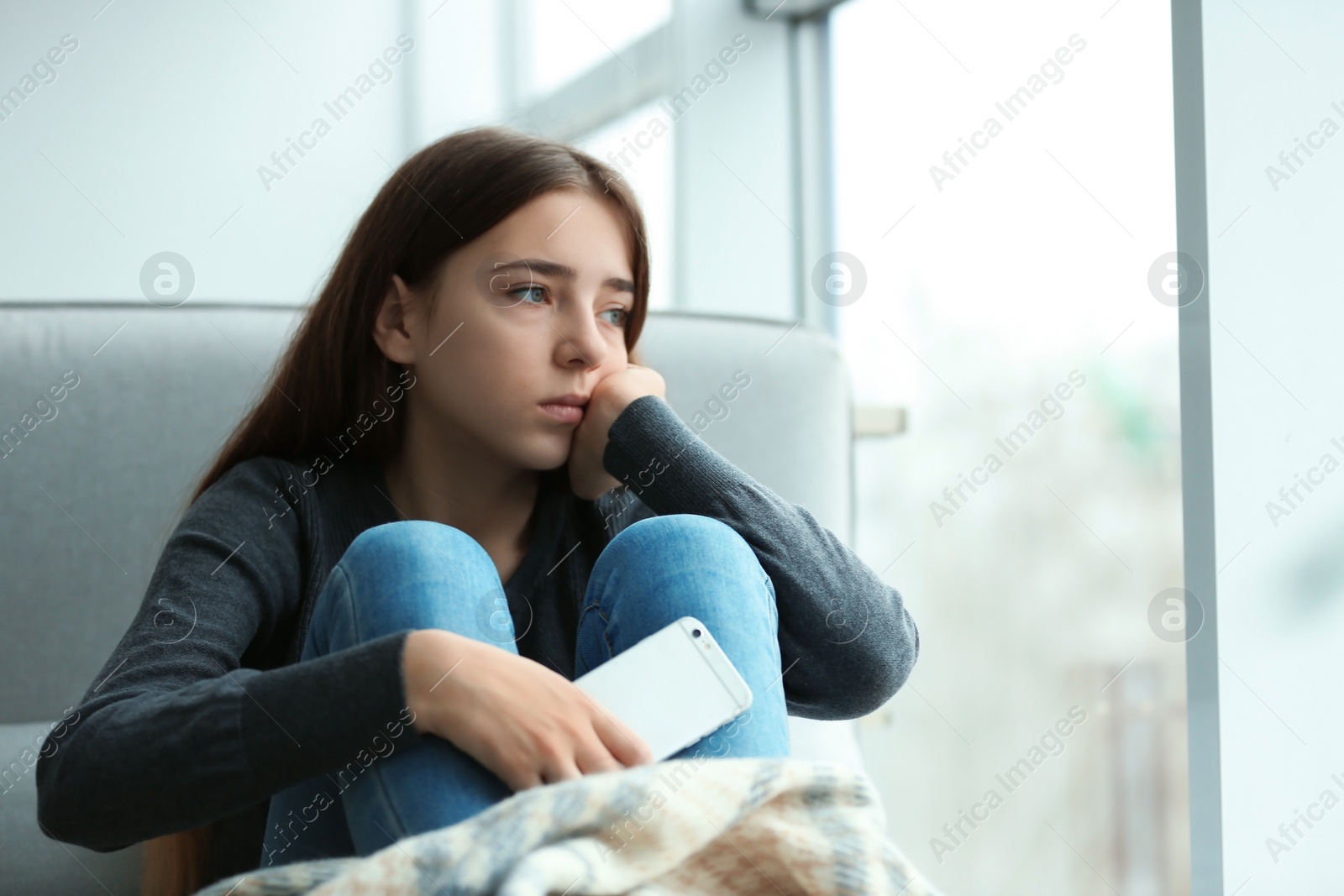 Photo of Upset teenage girl with smartphone sitting at window indoors. Space for text