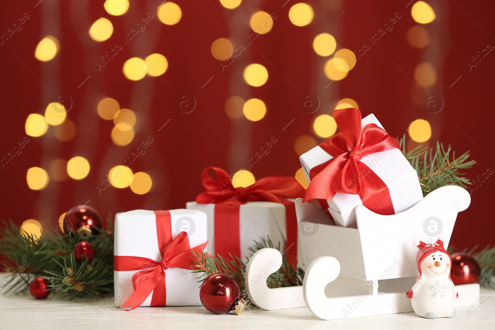 Photo of Decorative sleigh with Christmas gift boxes and decor on white table against blurred lights. Space for text