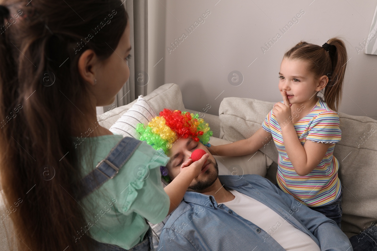 Photo of Cute little children putting clown nose and wig on father while he sleeping at home