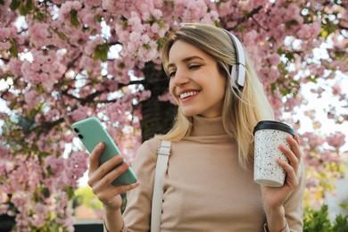 Photo of Happy woman with smartphone and coffee listening to audiobook outdoors on spring day