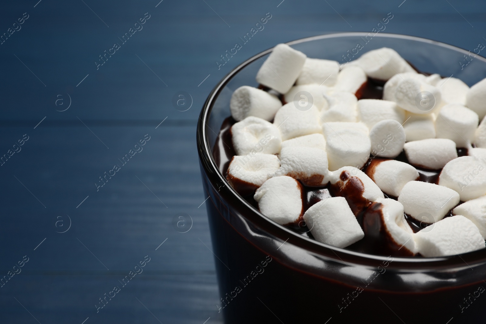 Photo of Glass cup of delicious hot chocolate with marshmallows on blue wooden table, closeup. Space for text
