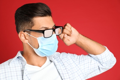 Man wiping foggy glasses caused by wearing medical mask on red background