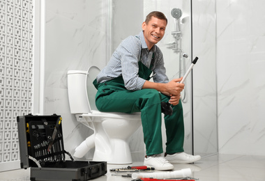 Photo of Professional plumber with plunger sitting on toilet bowl in bathroom