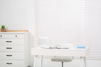 Photo of Beautiful office with laptop on white table, chair and chest of drawers. Doctor's workplace