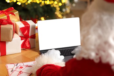 Photo of Santa Claus using laptop, closeup. Christmas gifts and letters on table indoors