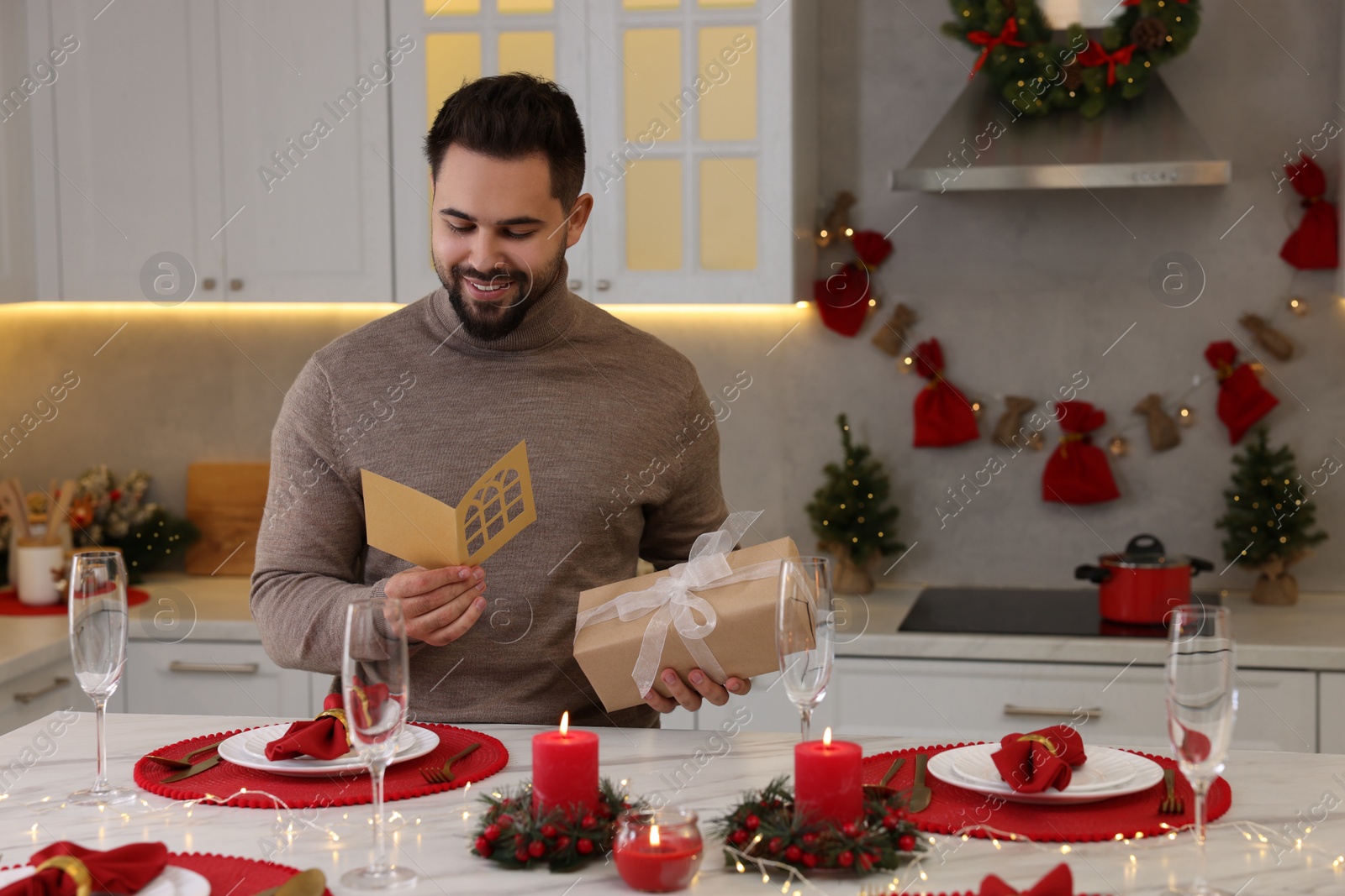 Photo of Happy young man with Christmas gift reading greeting card at table in kitchen