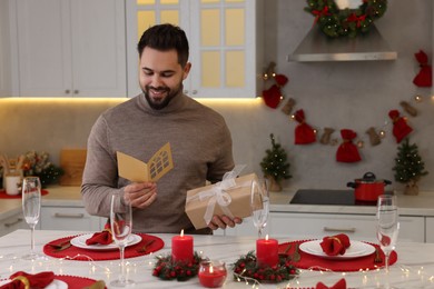 Happy young man with Christmas gift reading greeting card at table in kitchen