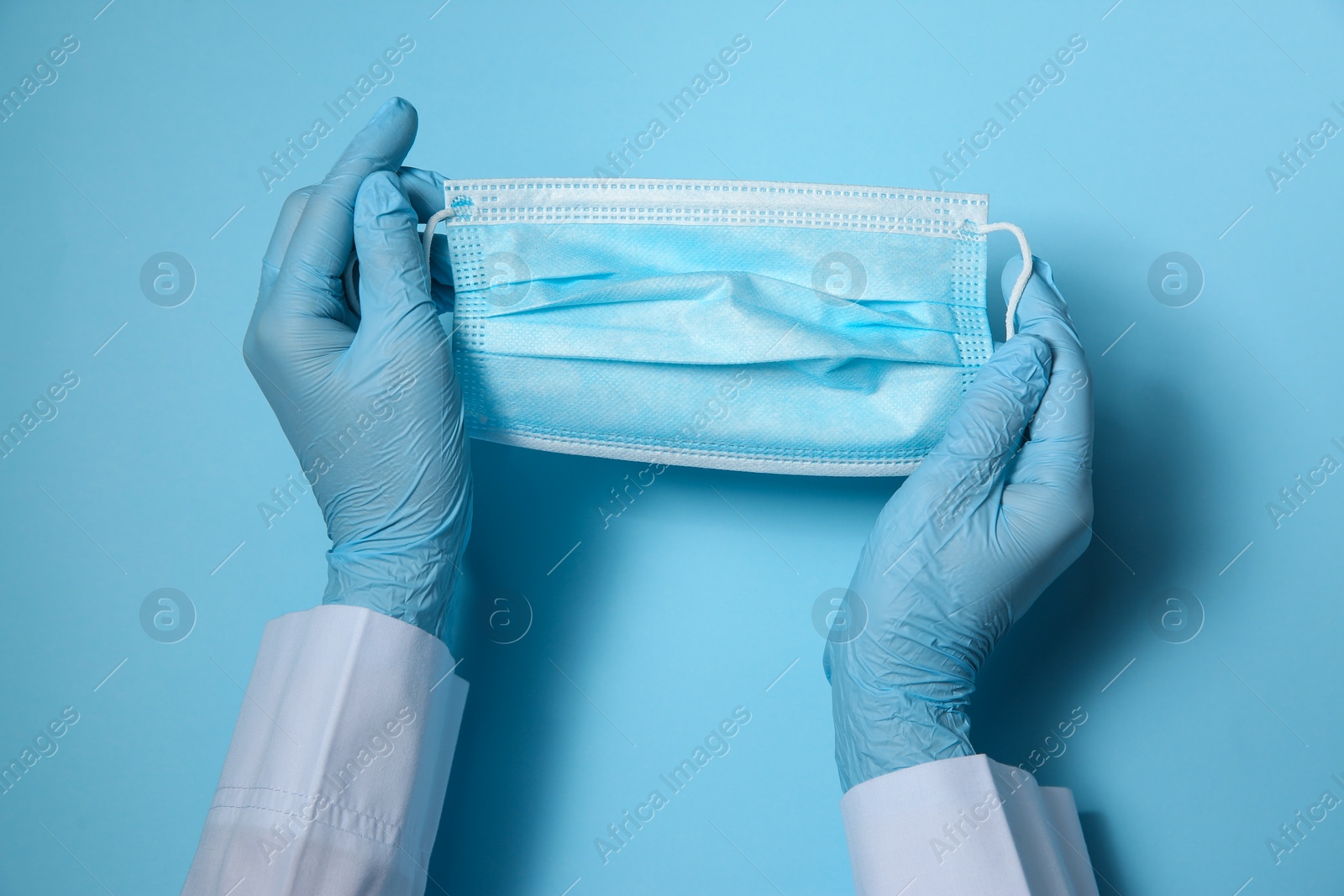 Photo of Doctor in latex gloves holding disposable face mask on light blue background, closeup. Protective measures during coronavirus quarantine