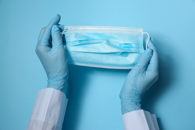 Photo of Doctor in latex gloves holding disposable face mask on light blue background, closeup. Protective measures during coronavirus quarantine