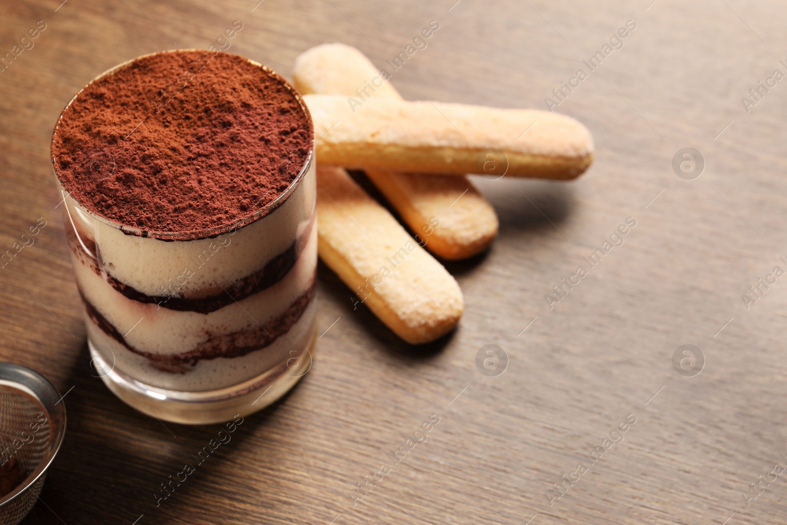 Photo of Delicious tiramisu in glass, biscuits and sieve with cocoa powder on wooden table, closeup. Space for text