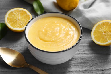 Delicious lemon curd in bowl, fresh citrus fruits and spoon on grey wooden table, closeup