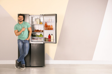 Photo of Happy young man near open refrigerator indoors, space for text