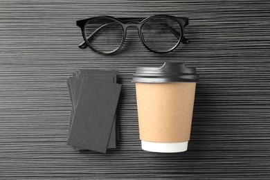 Blank black business cards, paper cup of coffee and glasses on wooden table, flat lay. Mockup for design