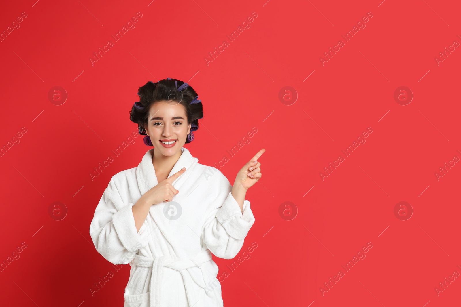 Photo of Happy young woman in bathrobe with hair curlers on red background, space for text