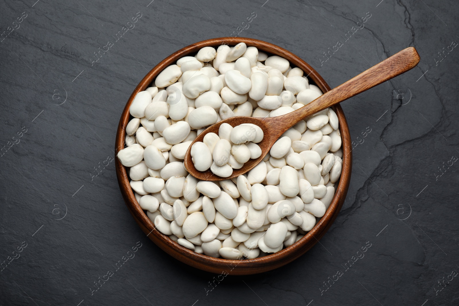 Photo of Raw white beans and spoon in bowl on black table, top view