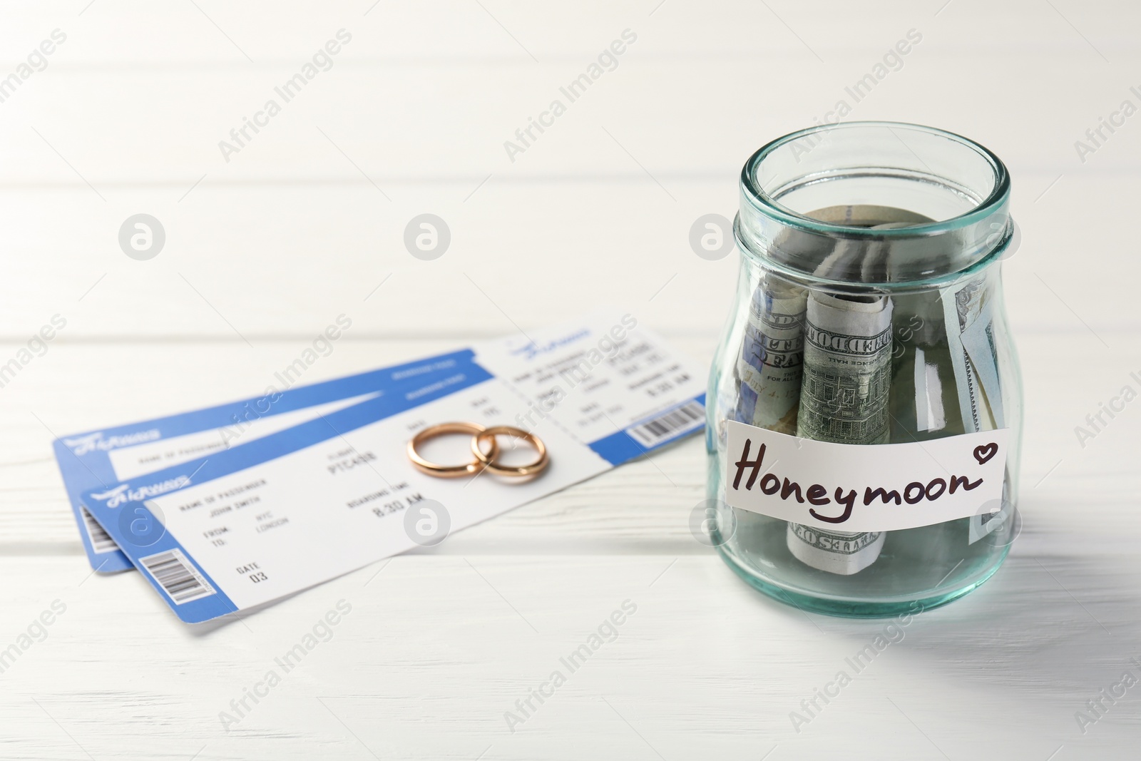 Photo of Glass jar with word Honeymoon, dollar banknotes, plane tickets and golden rings on white wooden table, closeup