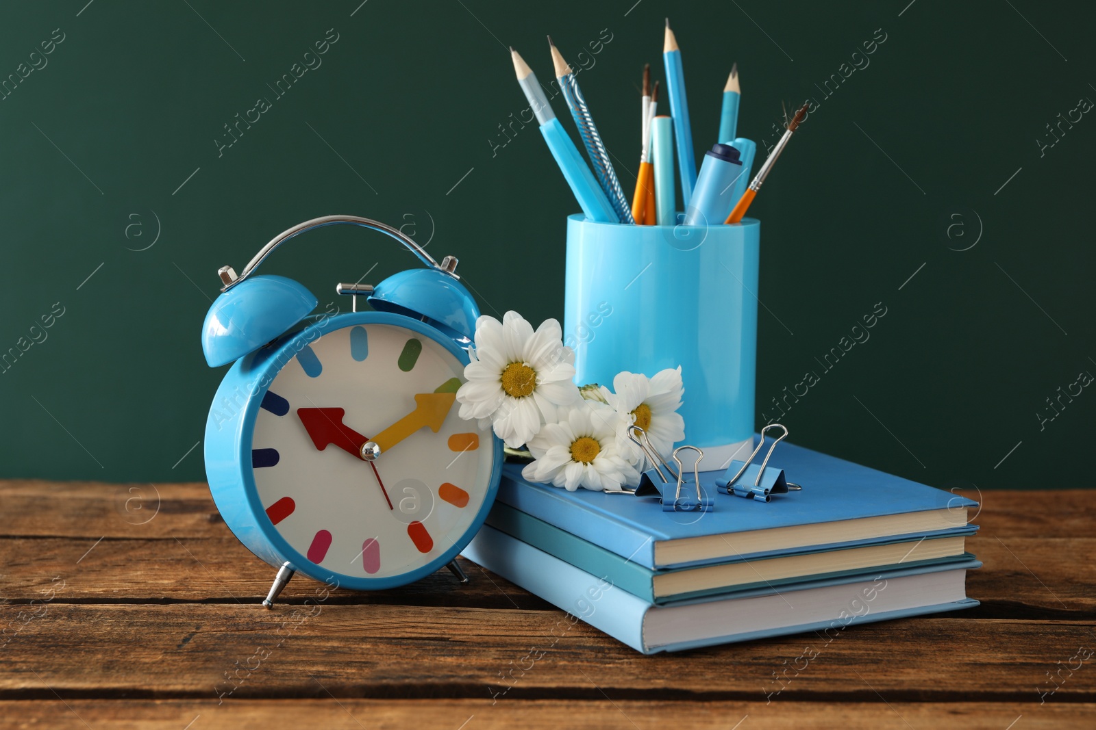 Photo of Alarm clock, flowers and set of stationery on wooden table near chalkboard. Teacher's Day