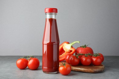 Photo of Bottle of tasty ketchup, tomatoes and pepper on grey table