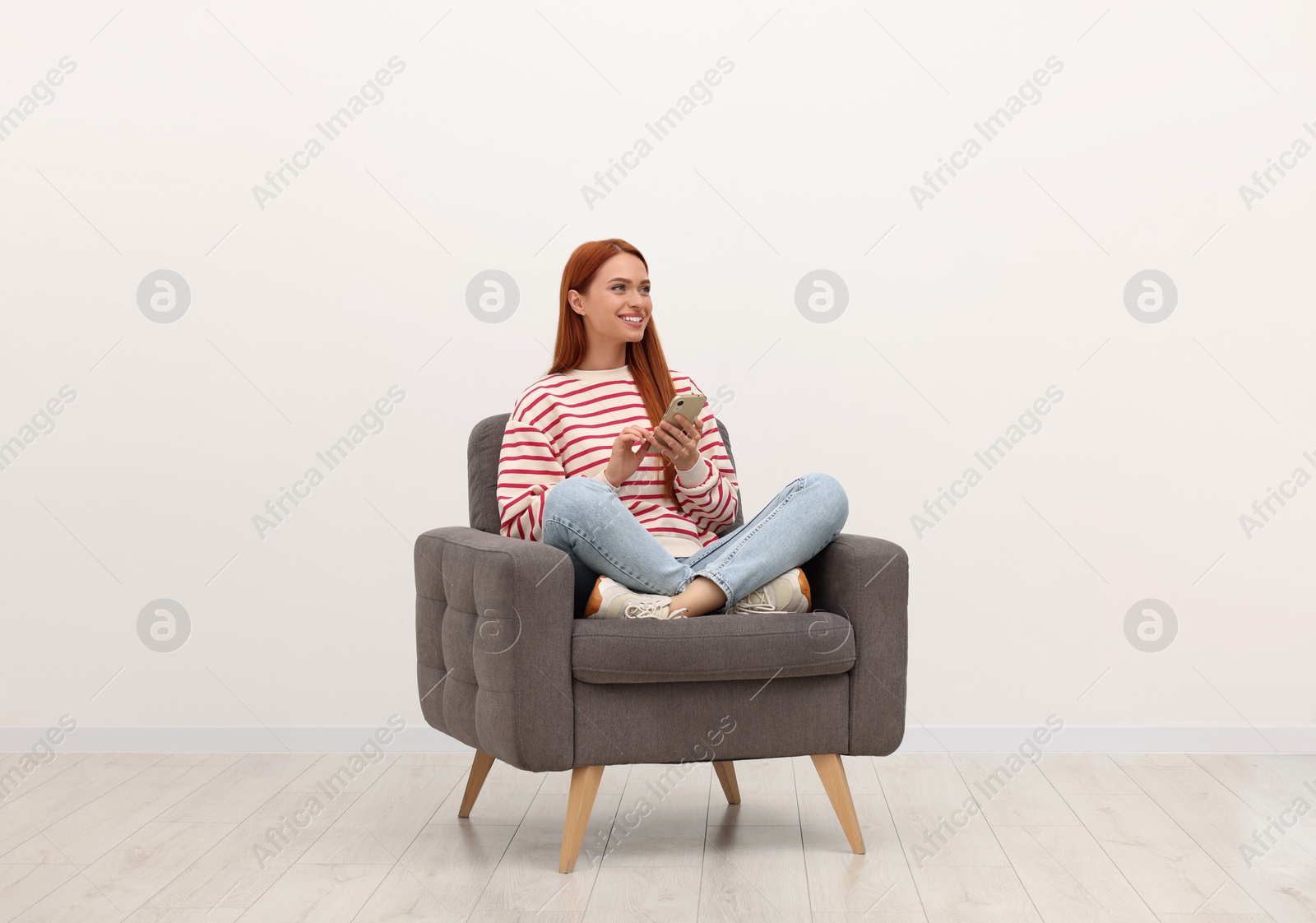 Photo of Happy young woman with smartphone sitting in armchair near white wall indoors