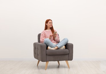 Happy young woman with smartphone sitting in armchair near white wall indoors