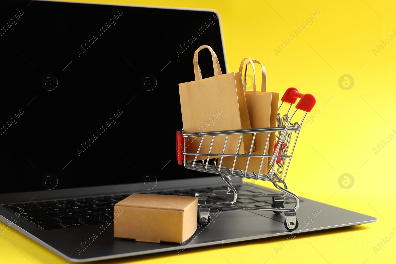 Photo of Online store. Laptop, mini shopping cart and purchases on yellow background