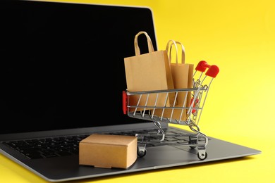 Photo of Online store. Laptop, mini shopping cart and purchases on yellow background