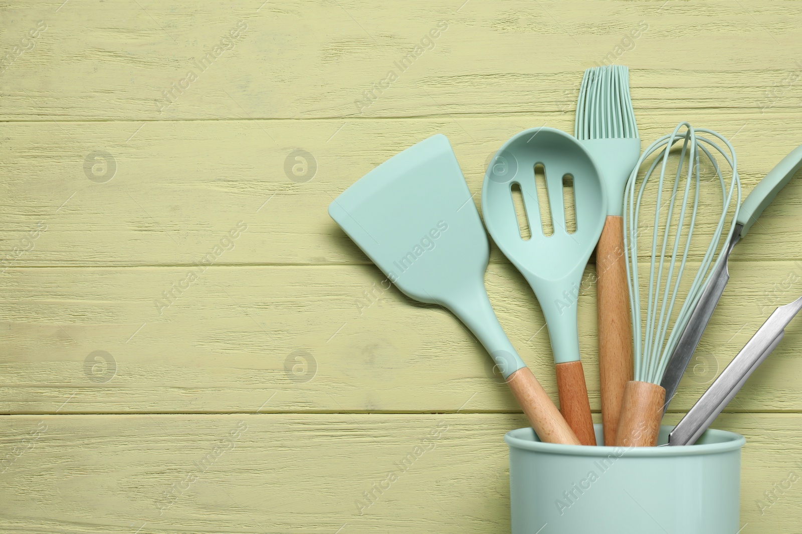 Photo of Set of kitchen utensils in holder on pale yellow wooden table, top view. Space for text