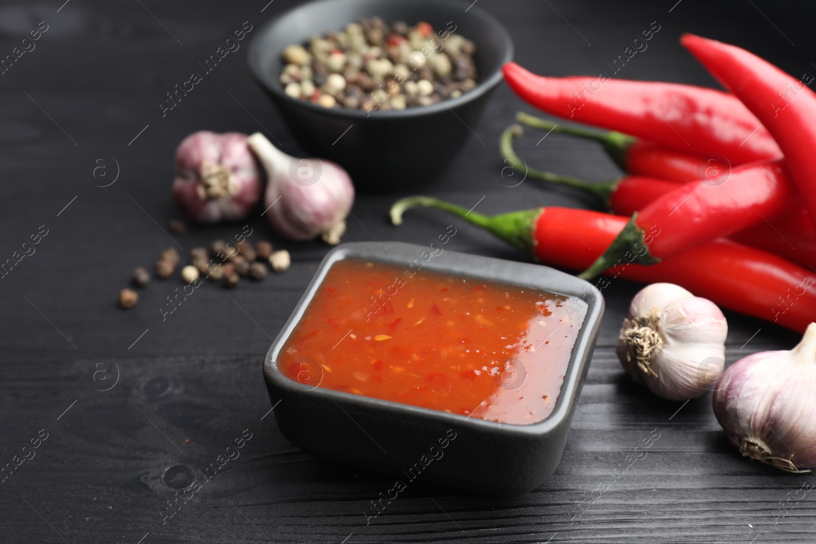 Photo of Spicy chili sauce, garlic, peppers and peppercorns on black wooden table, closeup