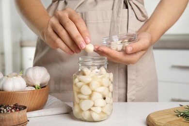 Woman taking clove of pickled garlic from jar at white wooden table in kitchen, closeup