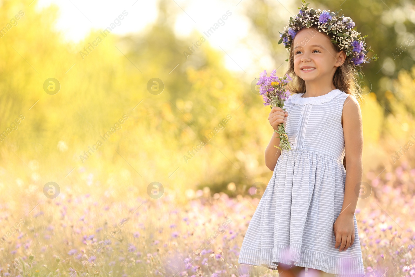 Photo of Cute little girl wearing beautiful wreath with bouquet of wildflowers outdoors, space for text. Child spending time in nature