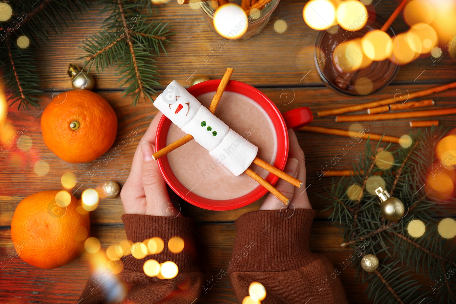 Image of Woman holding cup of hot drink with marshmallow snowman at wooden table, top view. Bokeh effect 