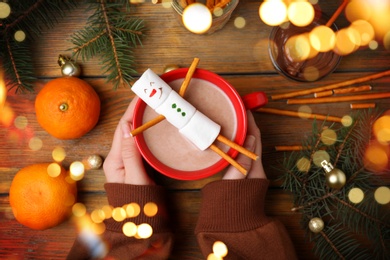 Woman holding cup of hot drink with marshmallow snowman at wooden table, top view. Bokeh effect 