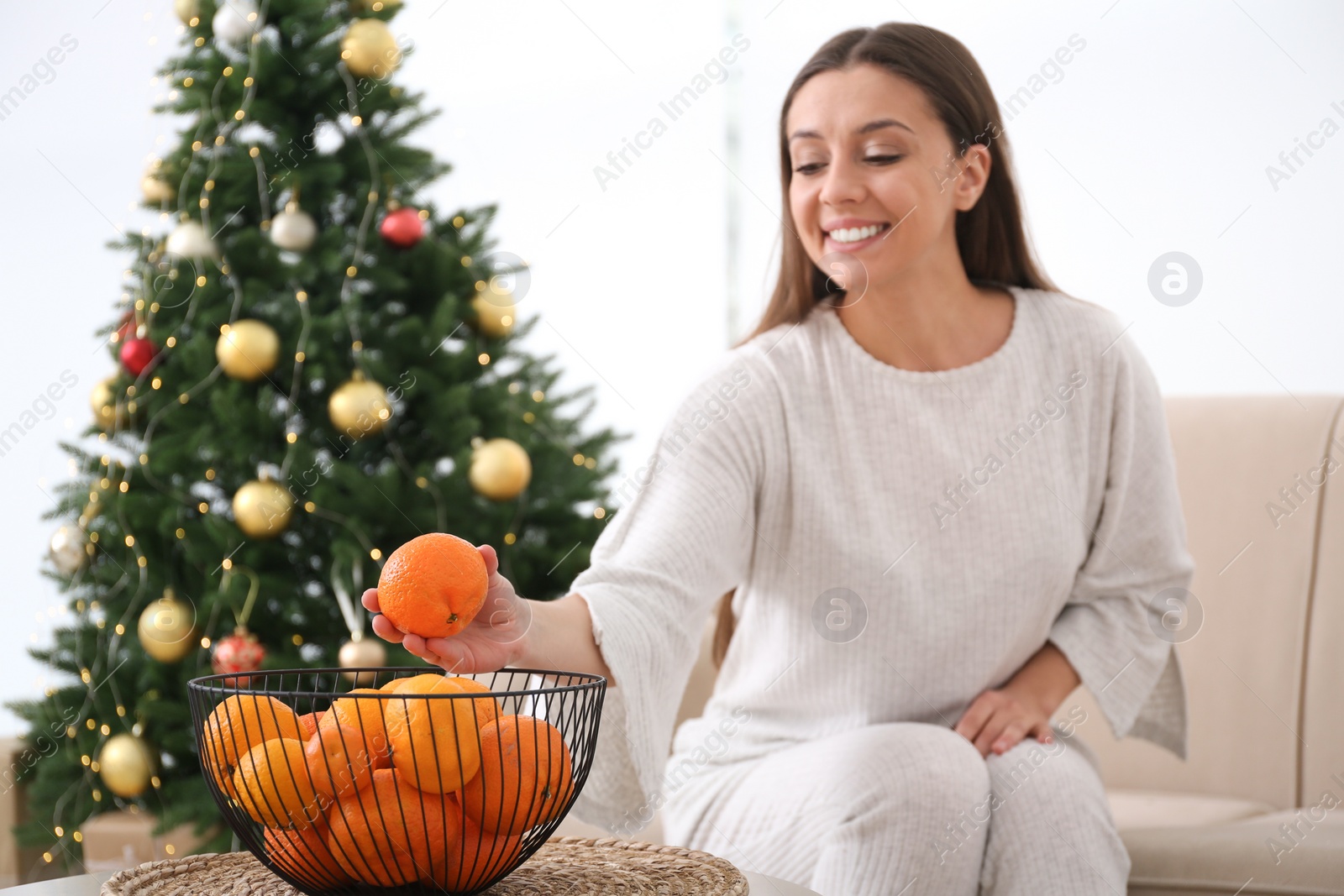 Photo of Woman taking tangerine from bowl near Christmas tree indoors