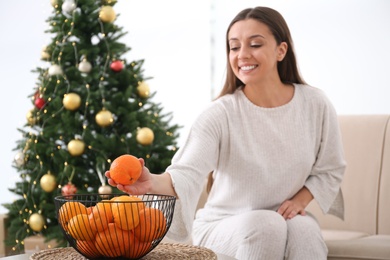 Woman taking tangerine from bowl near Christmas tree indoors