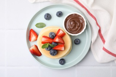 Photo of Delicious pancakes with strawberries, blueberries, mint and chocolate sauce on white tiled table, top view