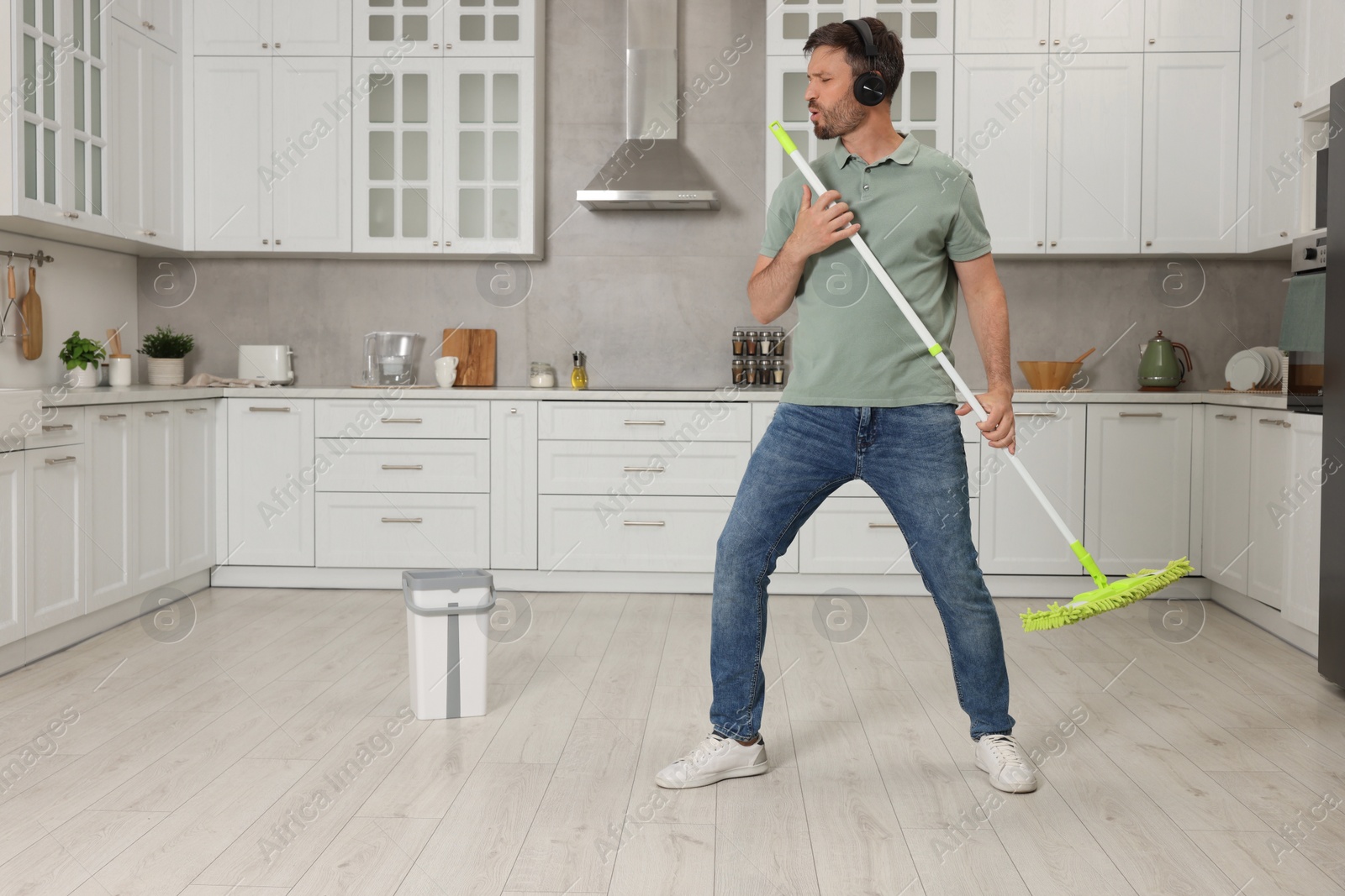 Photo of Enjoying cleaning. Happy man in headphones with mop singing while tidying up in kitchen