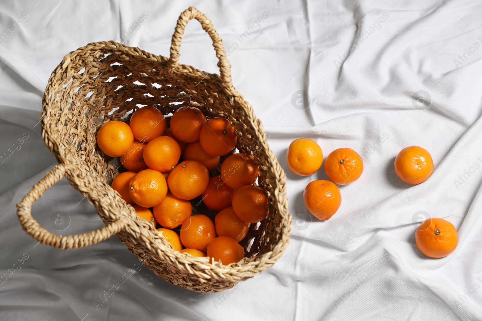 Photo of Stylish wicker bag with ripe tangerines on white bedsheet, flat lay