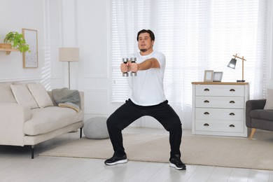 Photo of Overweight man doing squat exercise with dumbbells at home