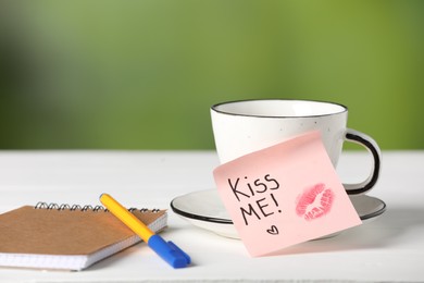 Photo of Sticky note with phrase Kiss Me, lipstick mark, cup of drink and notebook on white table against blurred background